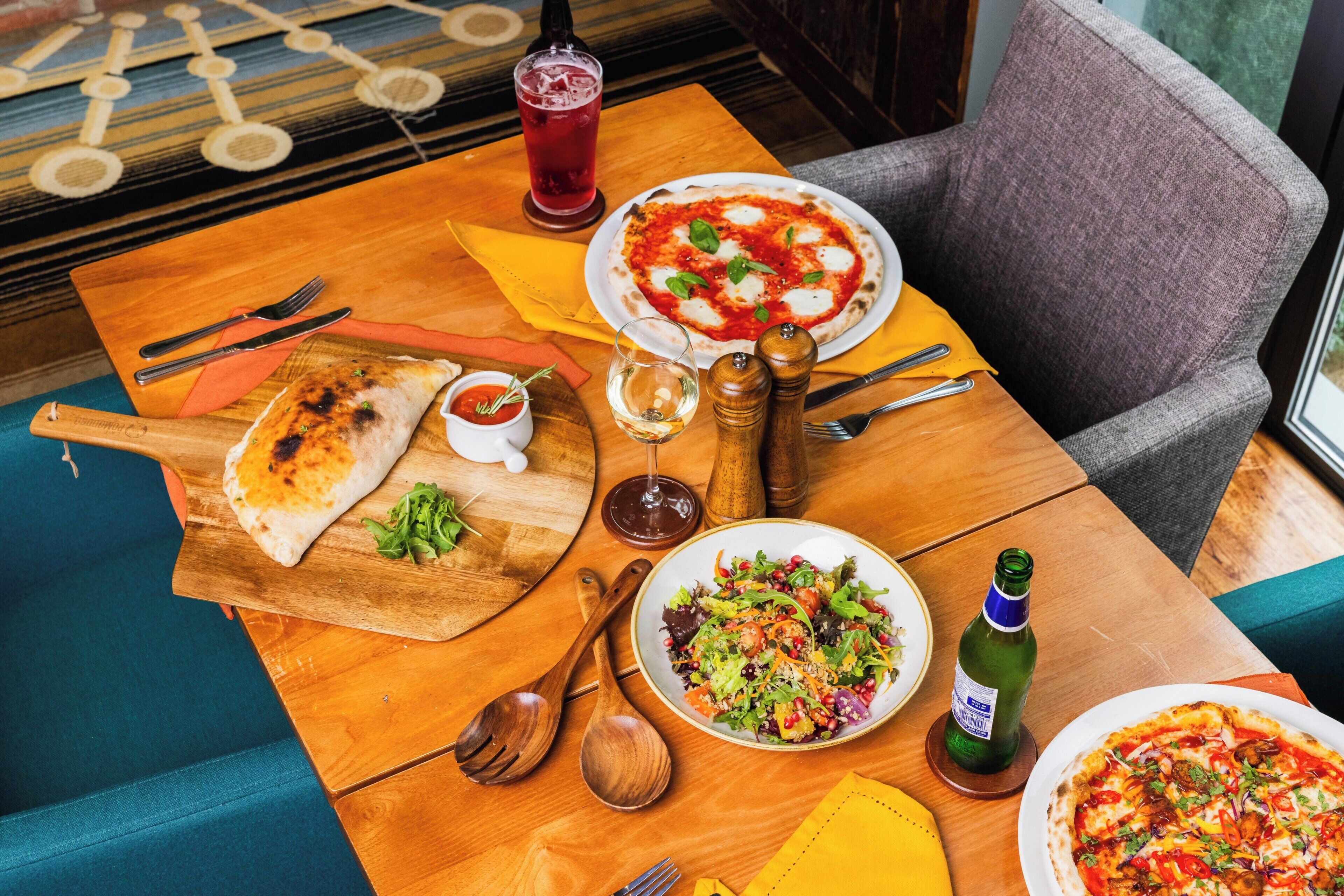 A wooden table with a variety of dishes including pizza, salad, and bread, along with cutlery and a glass of red drink.