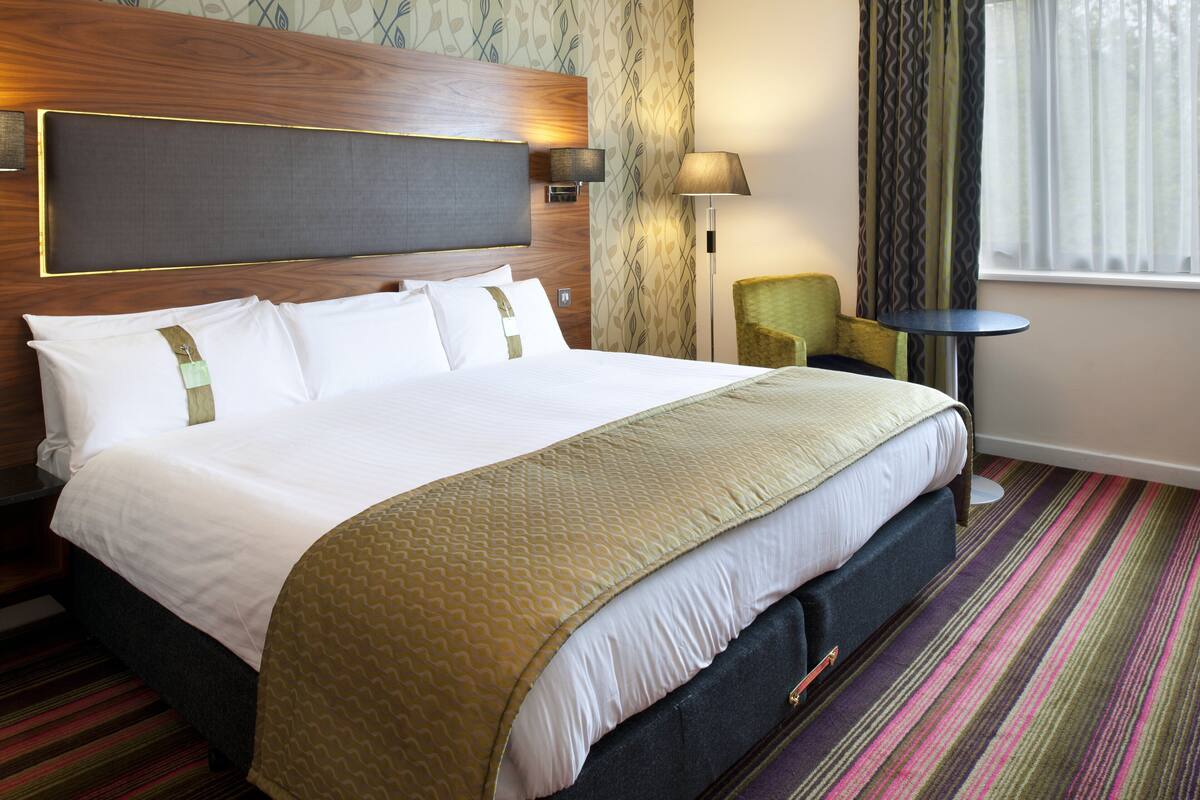 Habitación de hotel con una cama grande, cabecero, mesita de noche, lámpara, silla y ventana con cortinas.