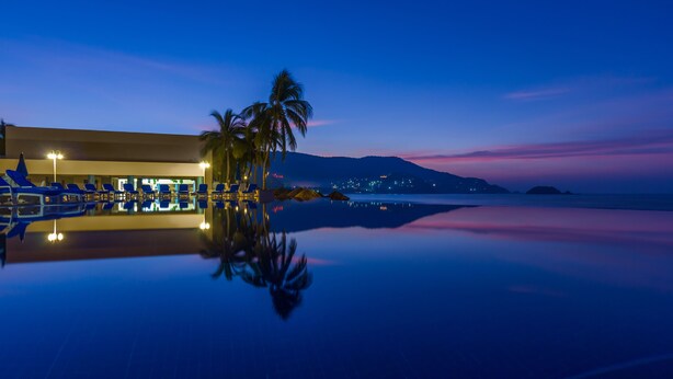 Zona junto a la piscina con sillones de descanso y una palmera, reflejando las luces y el cielo circundantes.