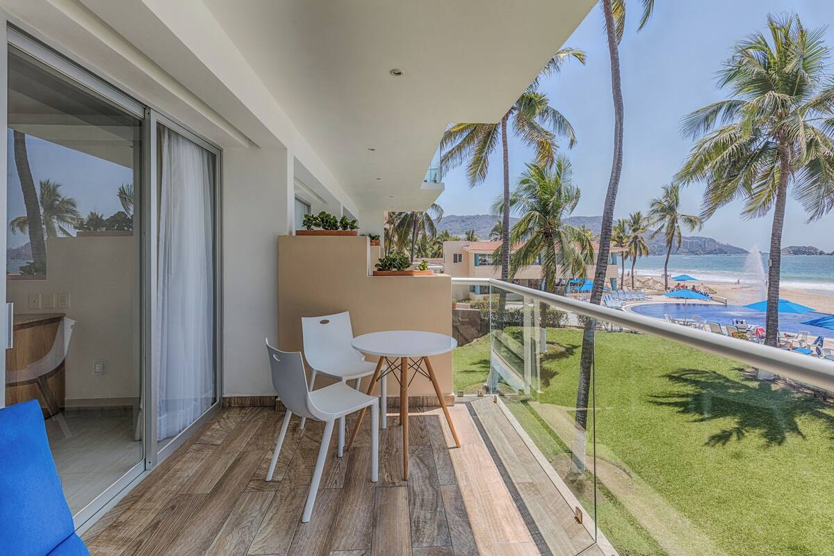 A balcony with a table and chairs, overlooking a beach with palm trees and a pool.
