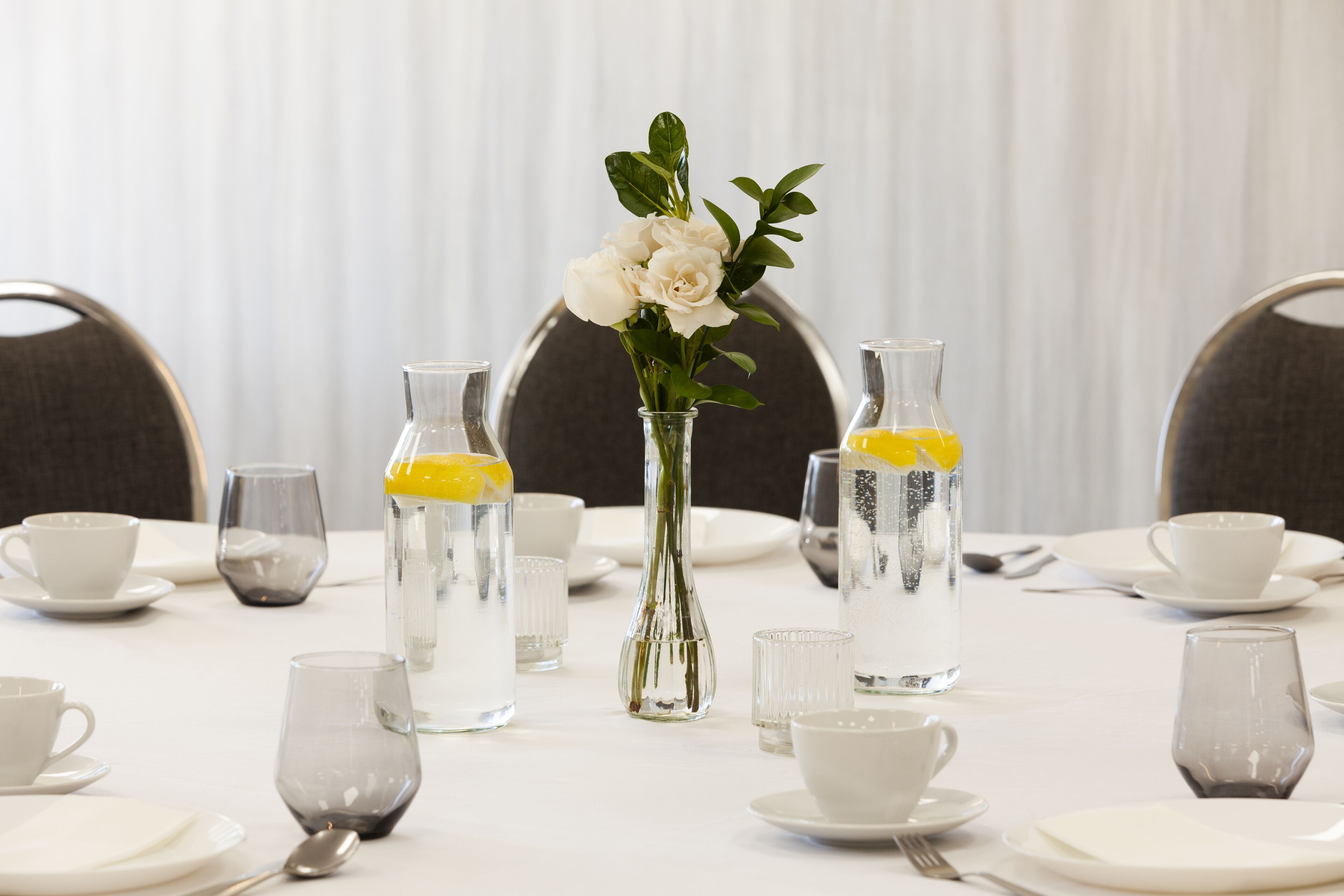 A dining table set with clear glass bottles, white cups, and a vase with white flowers.