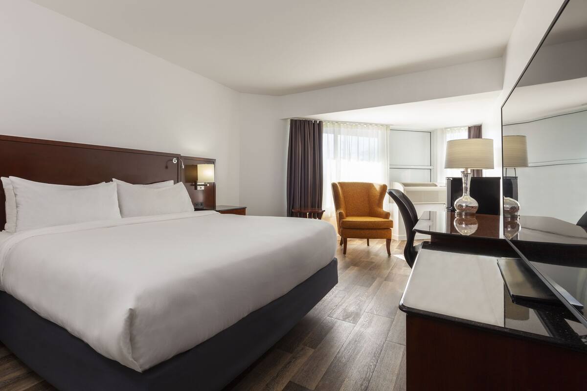A hotel room with a large bed, a desk with a chair, a mirror, and a window with curtains.