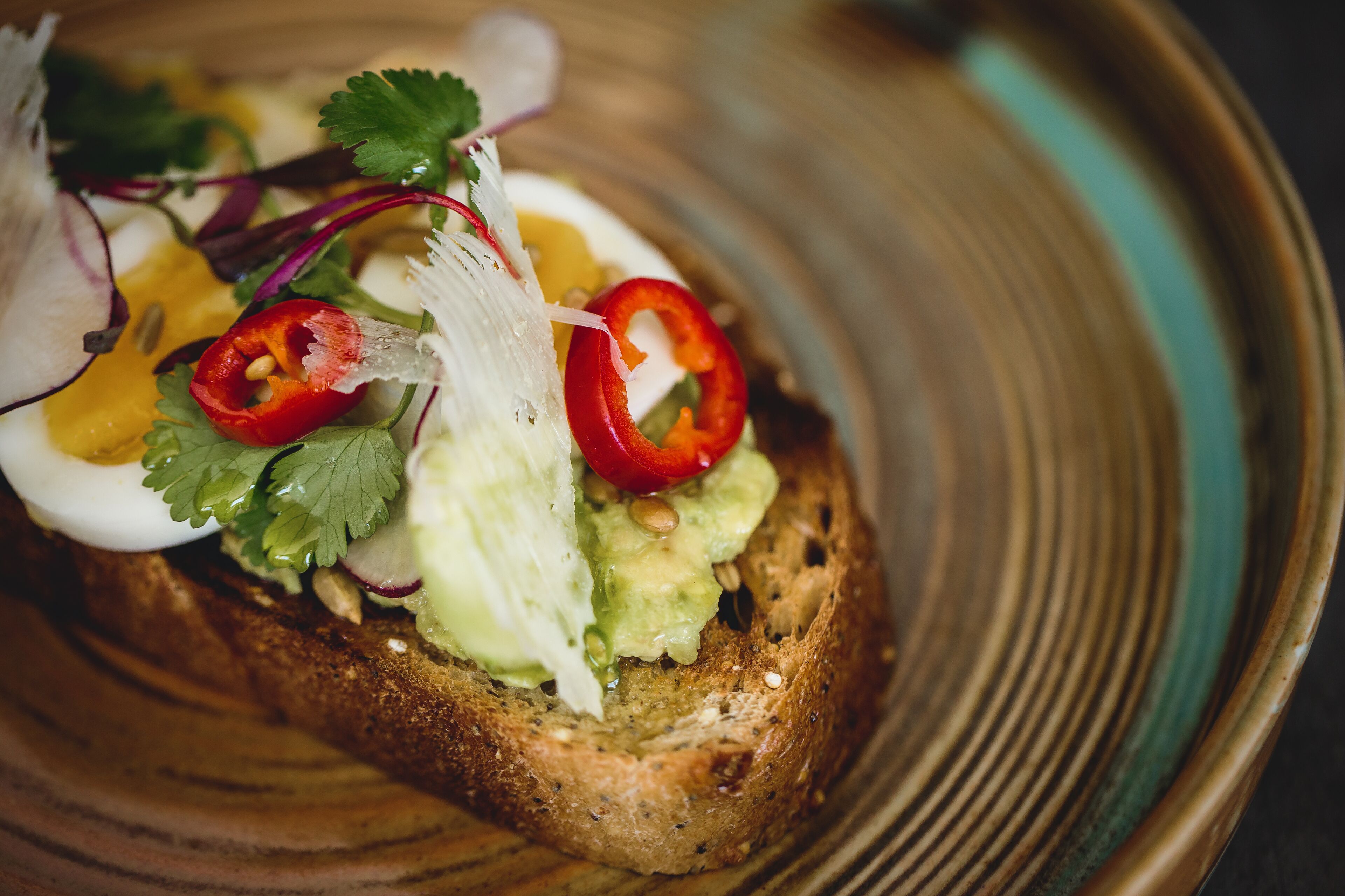 A slice of toast topped with avocado, red pepper, and a boiled egg, served on a ceramic plate.