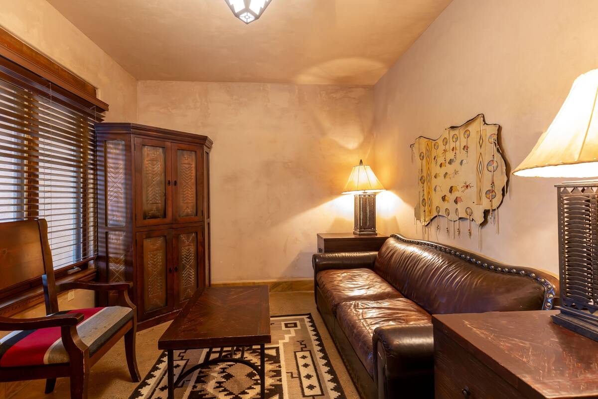 A living room with a brown leather sofa, wooden furniture, a patterned rug, and a wall-mounted lamp.