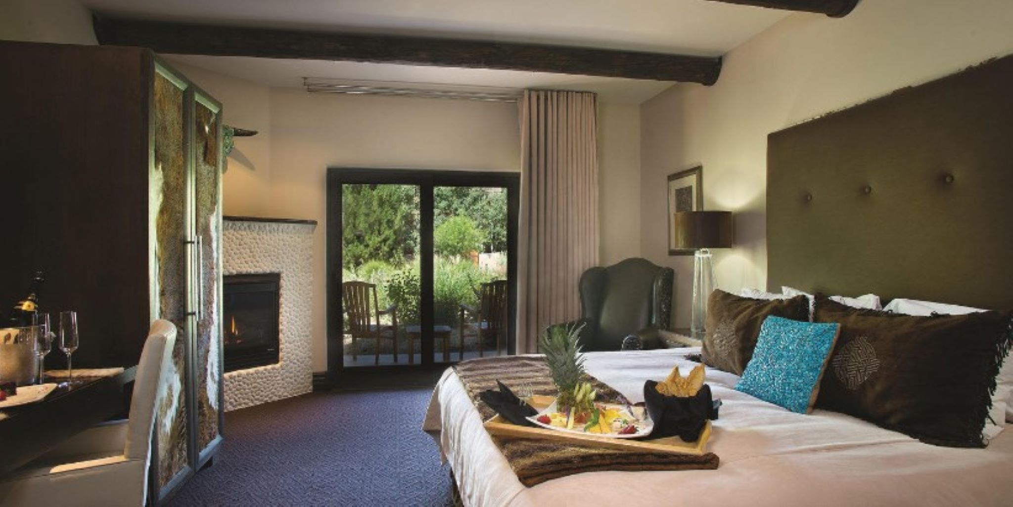 Habitación de hotel con chimenea, una cama con una bandeja de comida, una silla y vistas al exterior con vegetación.