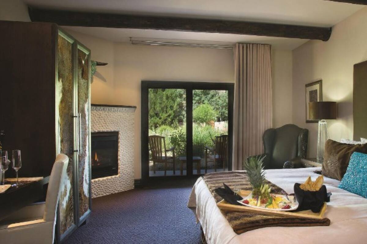 A hotel room with a fireplace, a bed with a tray of food, a chair, and a view of greenery outside.