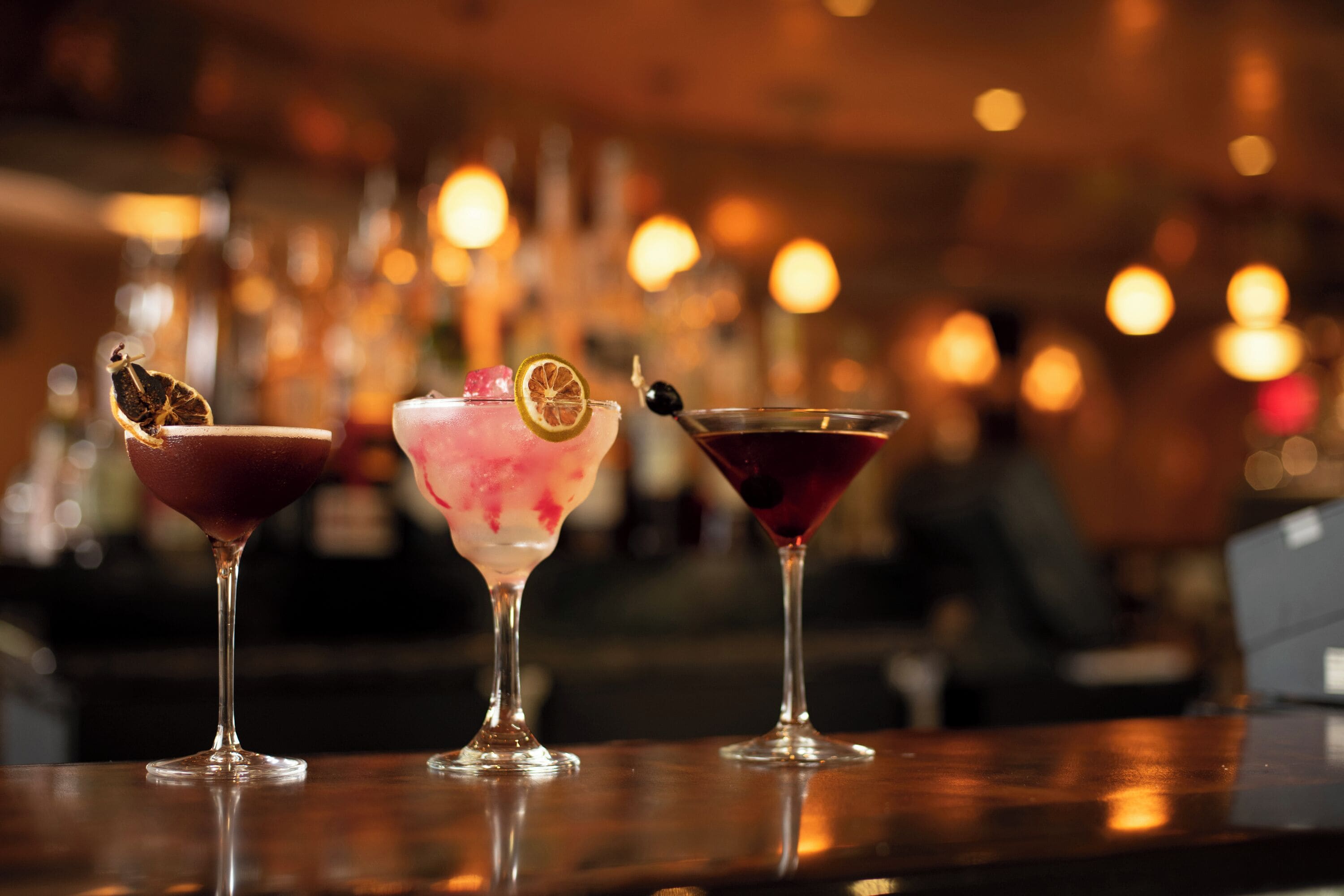 Three cocktails on a bar counter with a blurred background of lights.