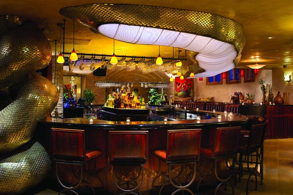 A bar area with a round bar, multiple stools, hanging lights, and a large decorative piece resembling a snake. - El Monte Sagrado Living Resort & Spa (Taos)