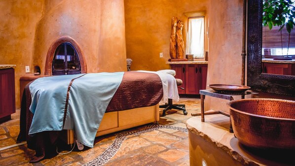 A spa room with a massage table, a stone floor, a large copper basin, and a wooden sculpture.