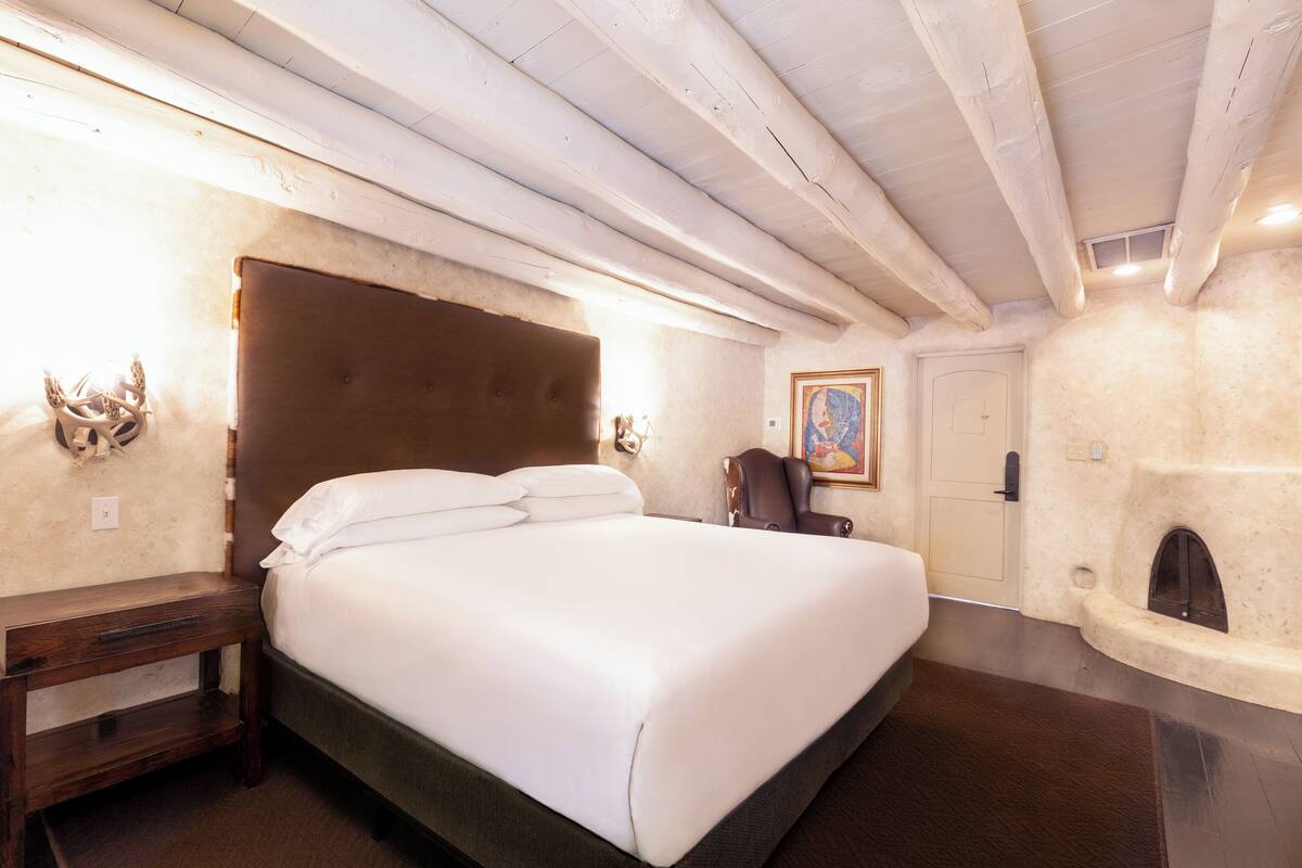 A bedroom with a large bed, a wooden nightstand, a fireplace, and a door with a painting.
