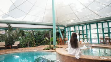 Una piscina interior con techo de cristal, rodeada de follaje y mobiliario de madera.