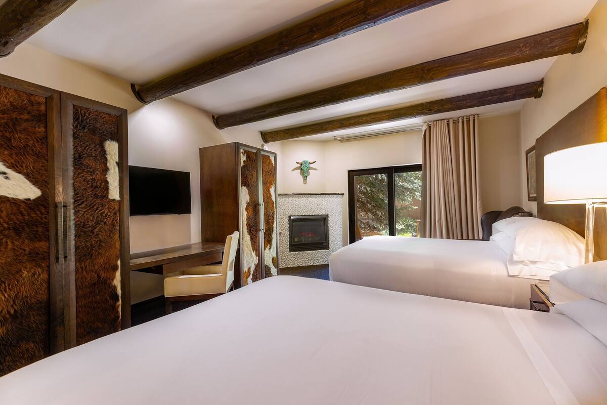A hotel room with two beds, a fireplace, wooden beams, a mounted TV, and a large window with curtains.