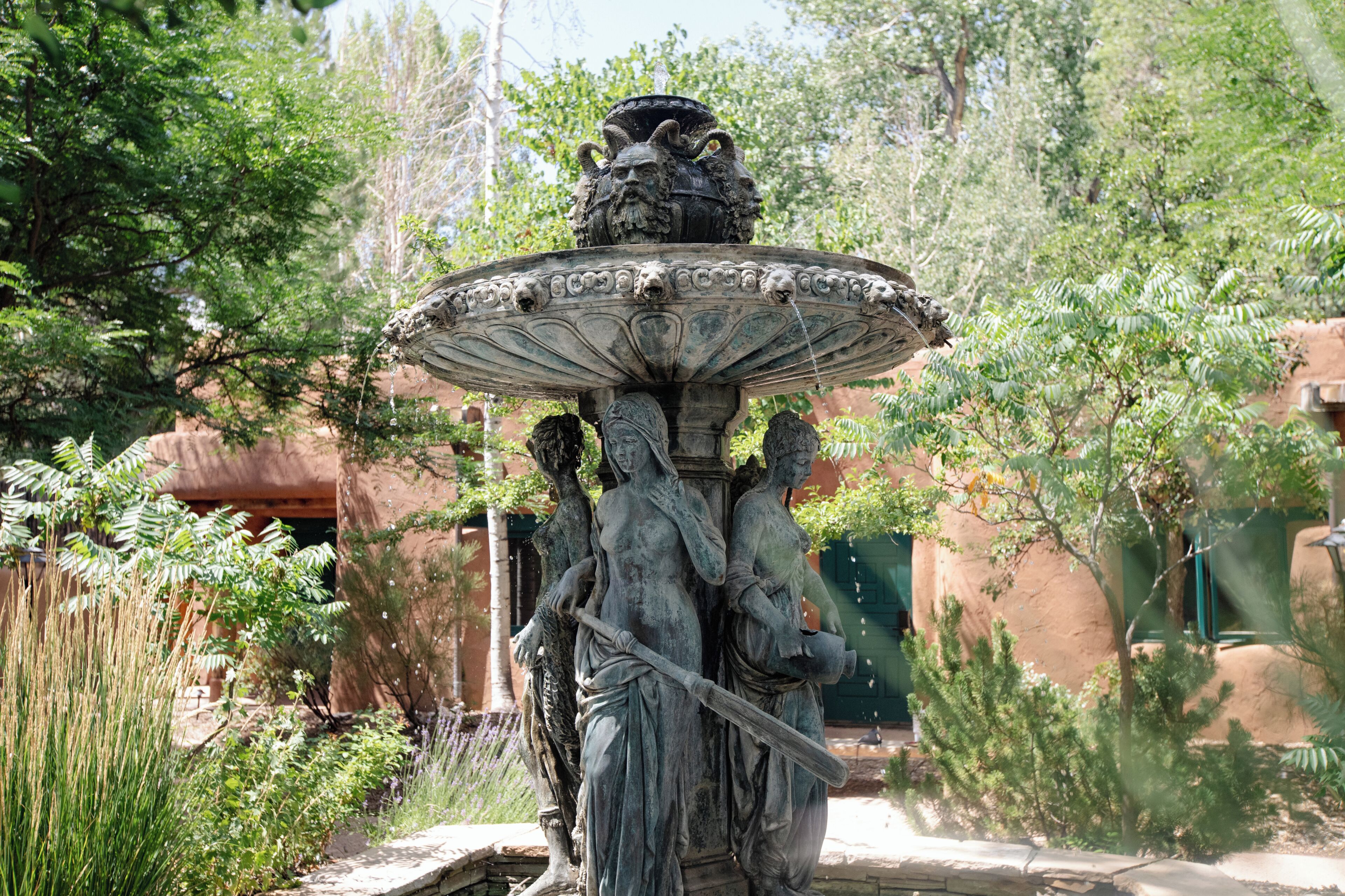 A fountain with a statue of a woman holding a staff, surrounded by greenery and a building in the background.