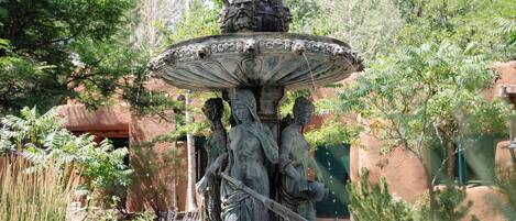 A fountain with a statue of a woman holding a staff, surrounded by greenery and a building in the background.