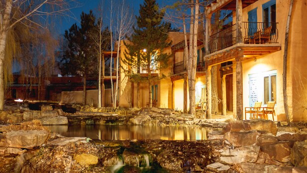 A lakeside lodge with a stone bridge, a waterfall, and a wooden deck overlooking the water.
