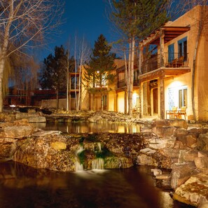 A lakeside lodge with a stone bridge, a waterfall, and a wooden deck overlooking the water.