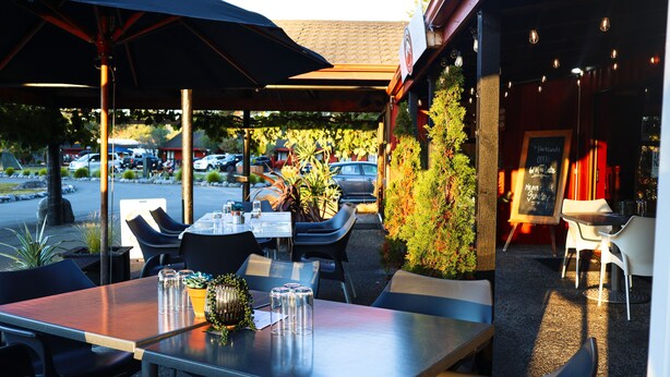 An outdoor dining area with tables and chairs, umbrellas, and a view of parked cars and greenery. - Parklands Motorlodge (Turangi)