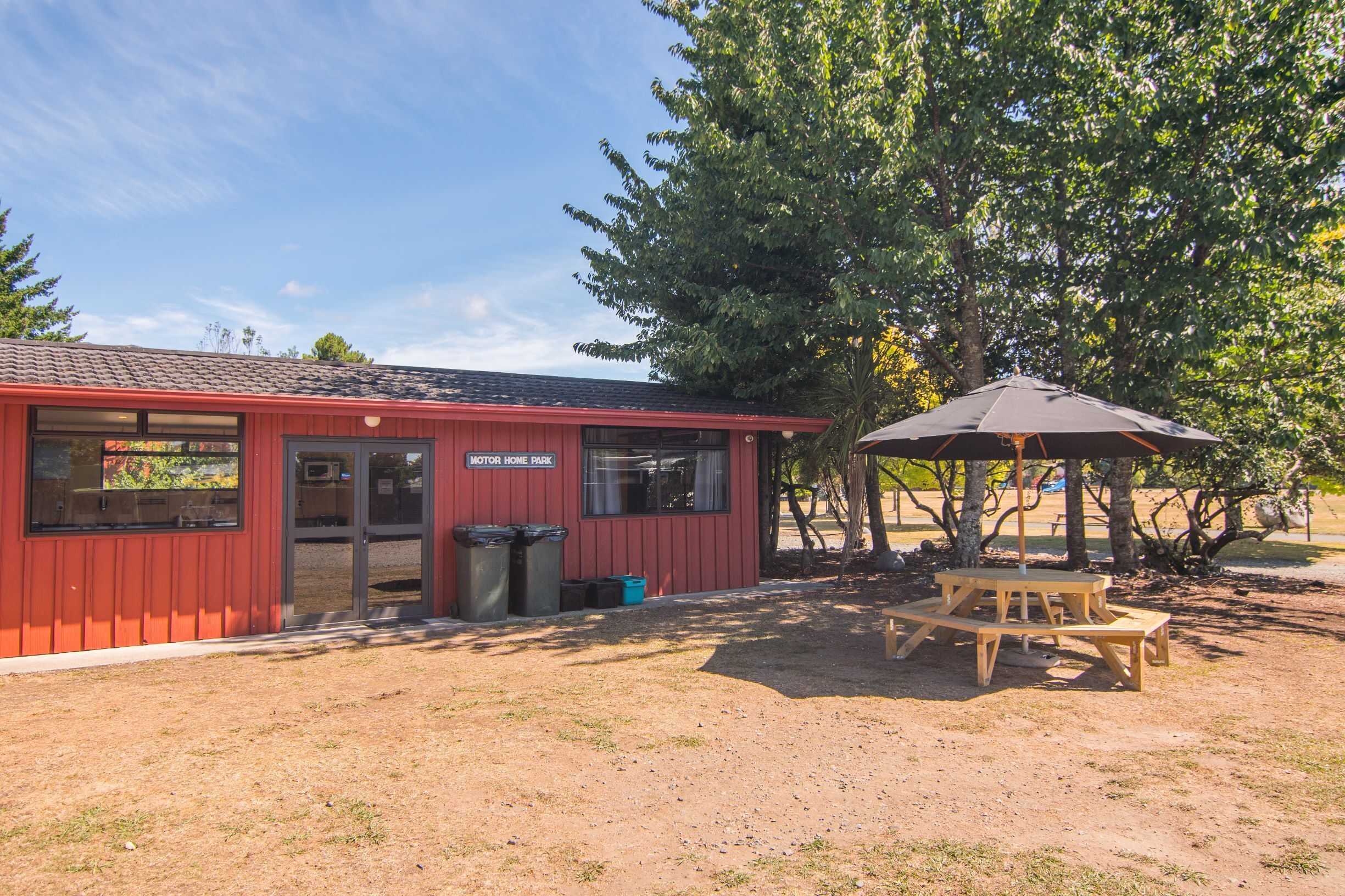 A red building labeled "Motor Home Park" with a picnic table and umbrella, surrounded by trees.