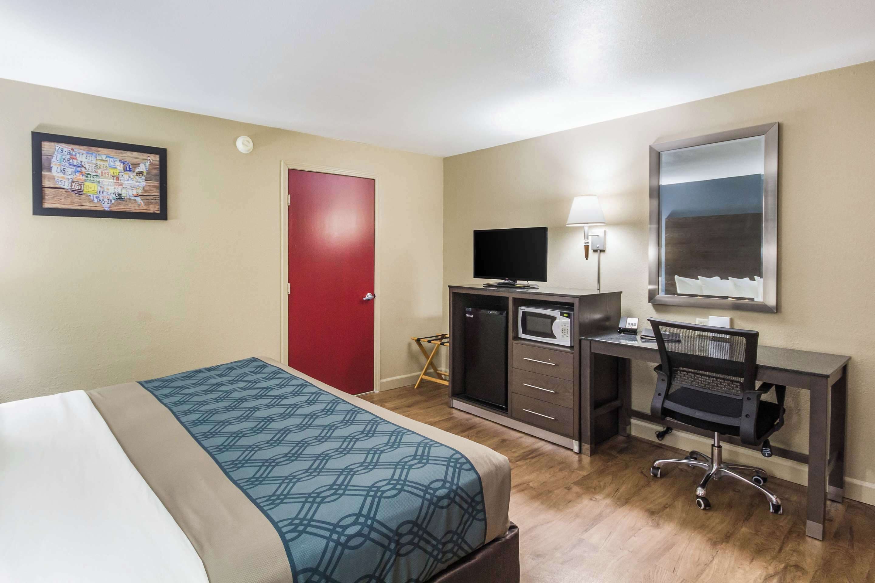 A hotel room with a bed, a desk with a computer, a television, and a red door.