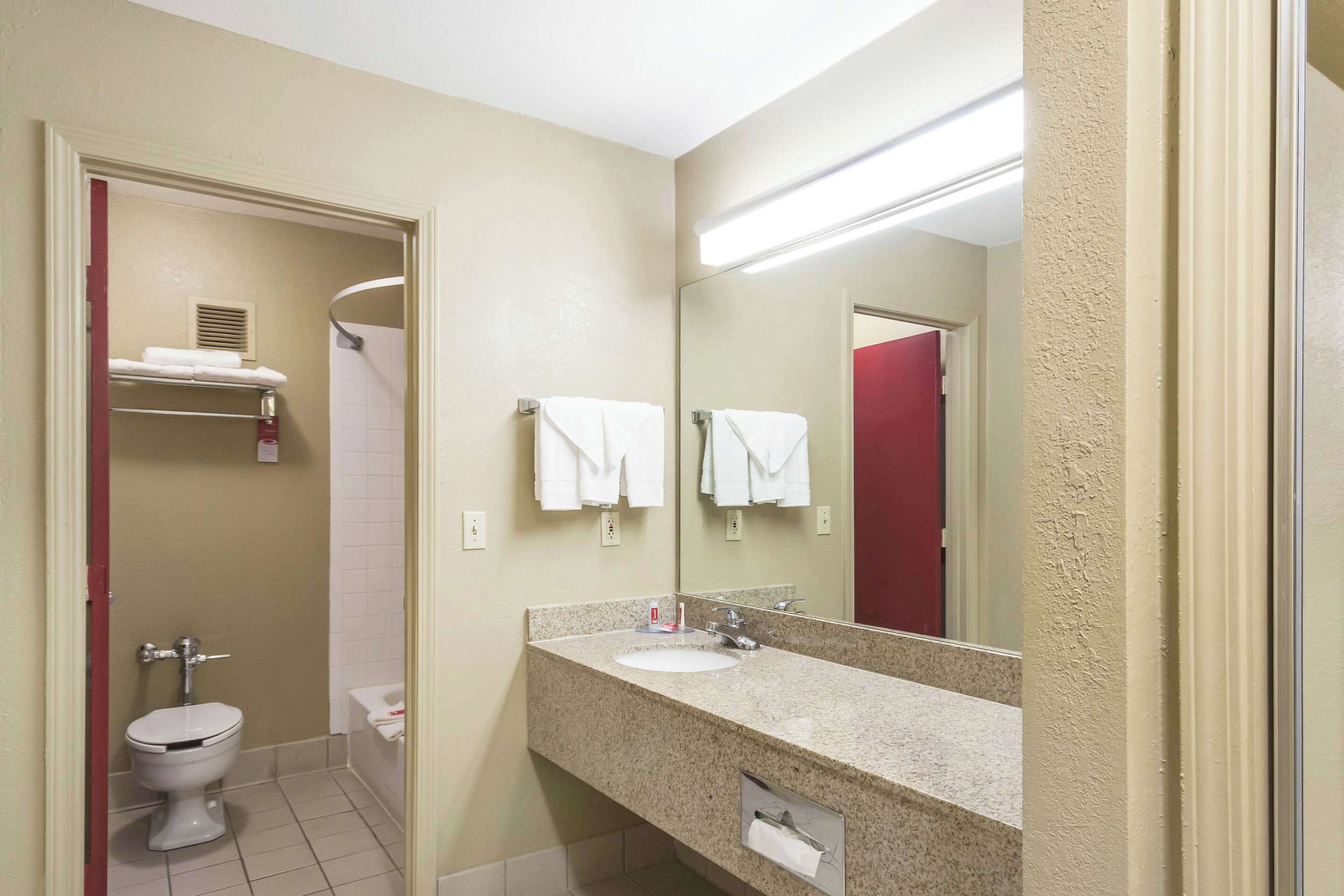 A bathroom with a toilet, sink, and mirror. There is a towel rack with towels and a shower visible in the background.