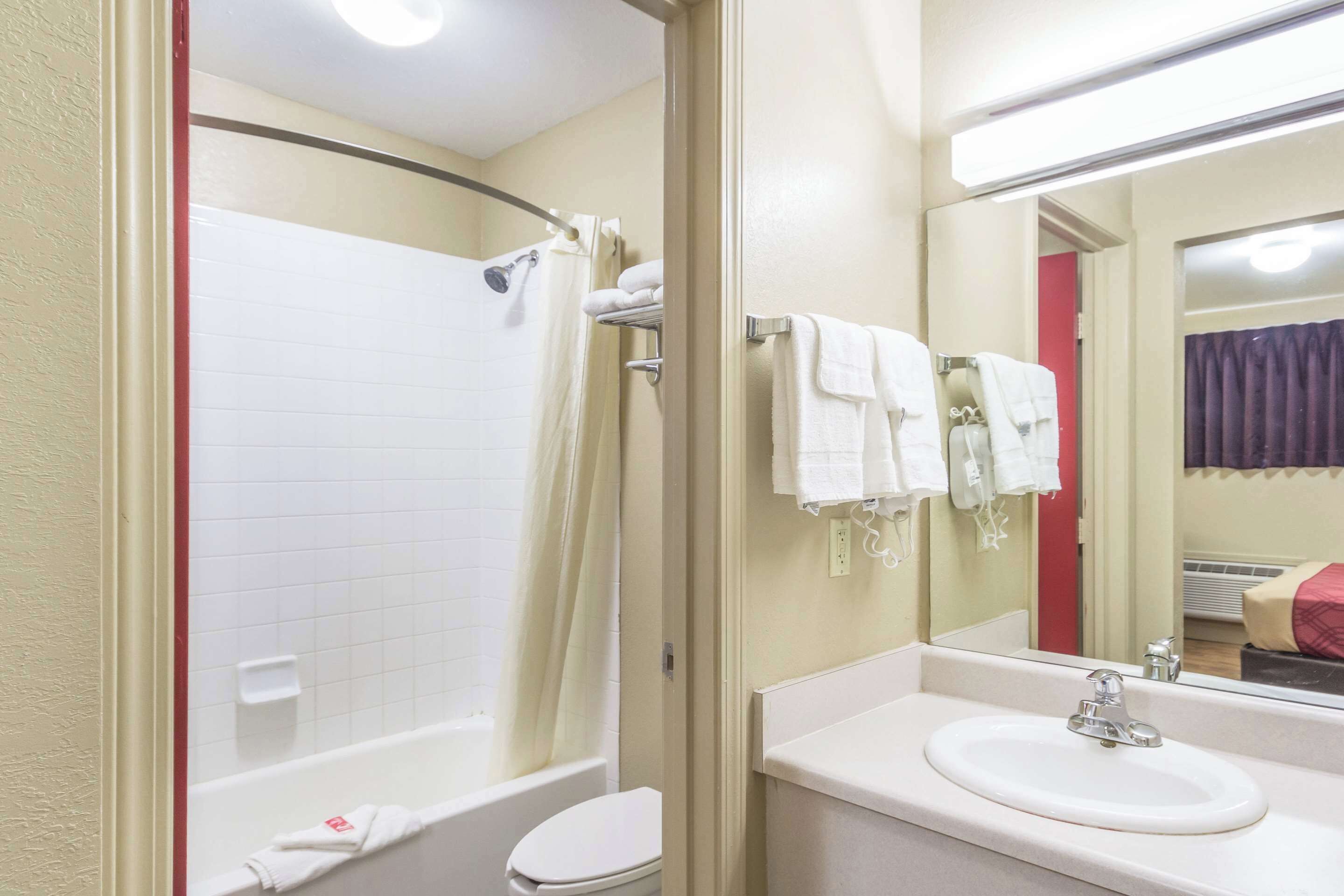 Un baño con bañera, inodoro, lavabo y un espejo que refleja una cama con una colcha roja.