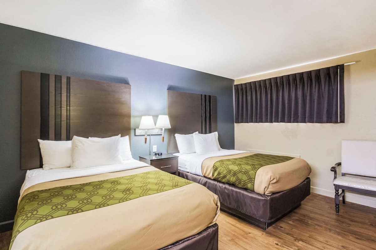 A hotel room with two beds, a nightstand with lamps, a chair, and a large window with curtains.