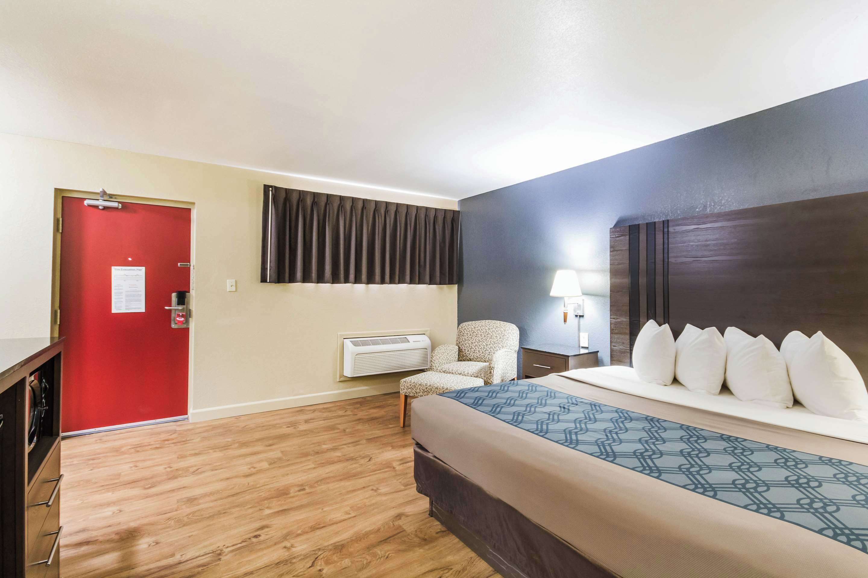 A hotel room with a bed, a chair, a desk, and a red door.