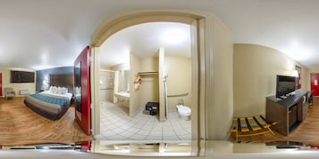 Una vista panorámica de una habitación de hotel con cama, un escritorio con televisor, un baño con inodoro y lavabo, y una zona de ducha.