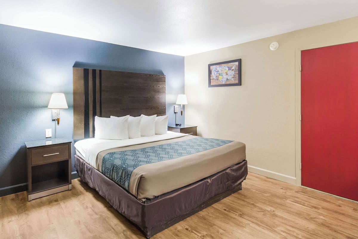 A neatly arranged hotel room with a bed, bedside lamps, a nightstand, a red door, and a wall-mounted artwork.