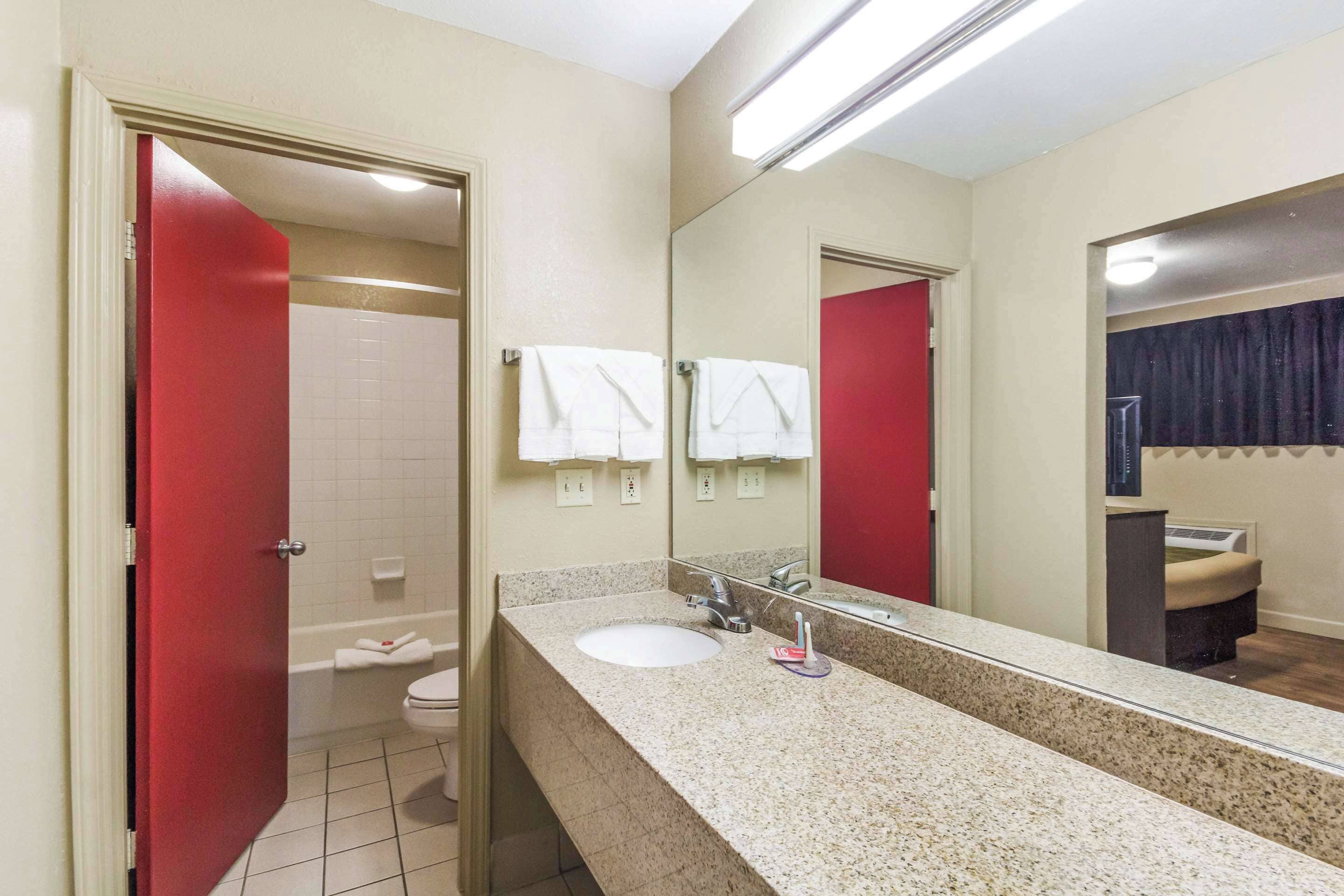 A hotel bathroom with a granite countertop sink, a mirror, and a red door leading to the shower.