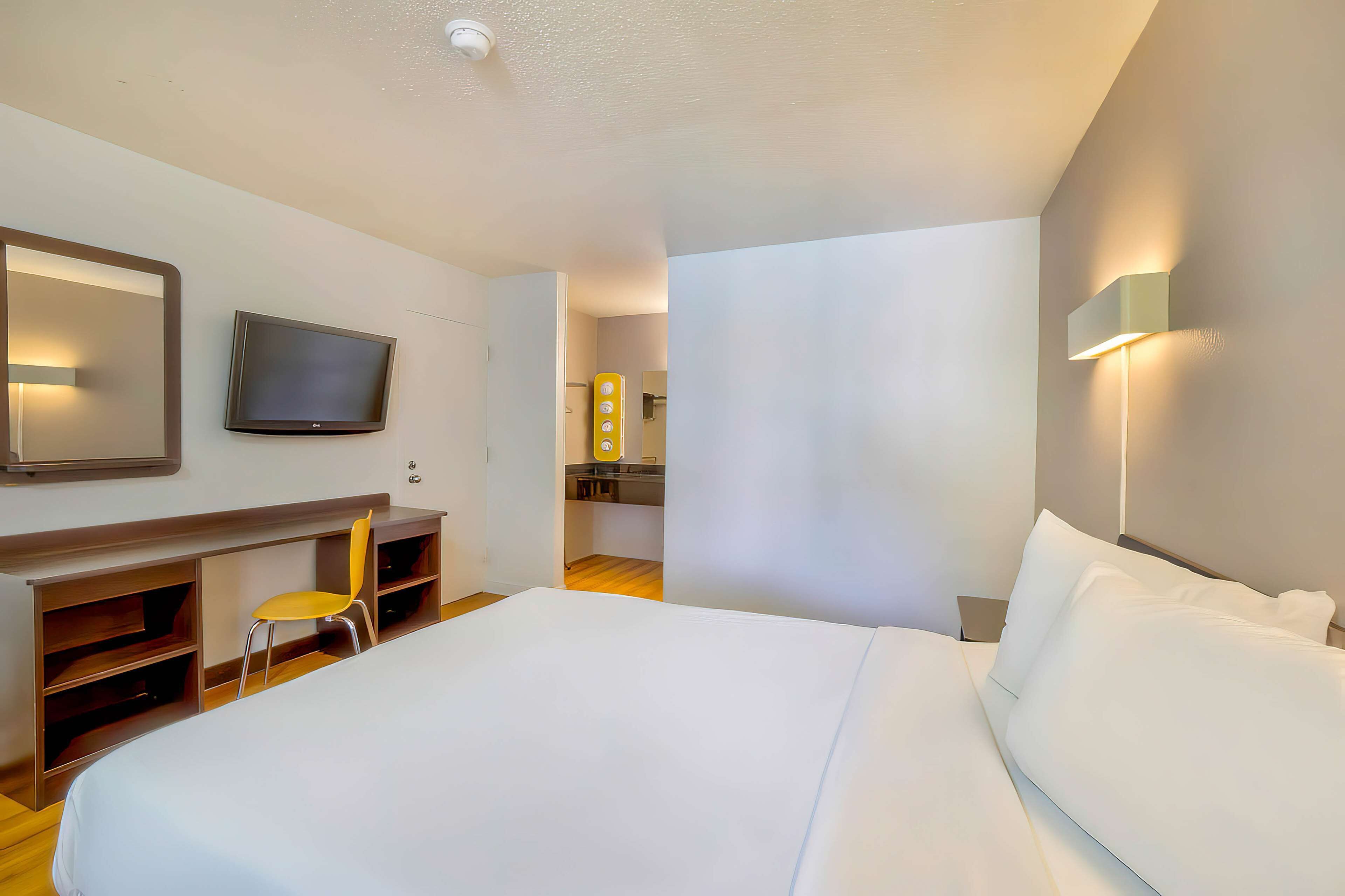 A hotel room with a large bed, a TV mounted on the wall, a desk with a chair, and a small kitchenette in the background.