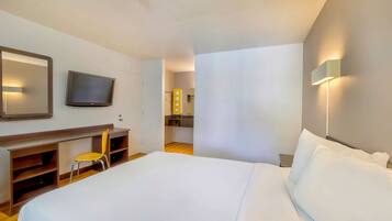 A hotel room with a large bed, a TV mounted on the wall, a desk with a chair, and a small kitchenette in the background.