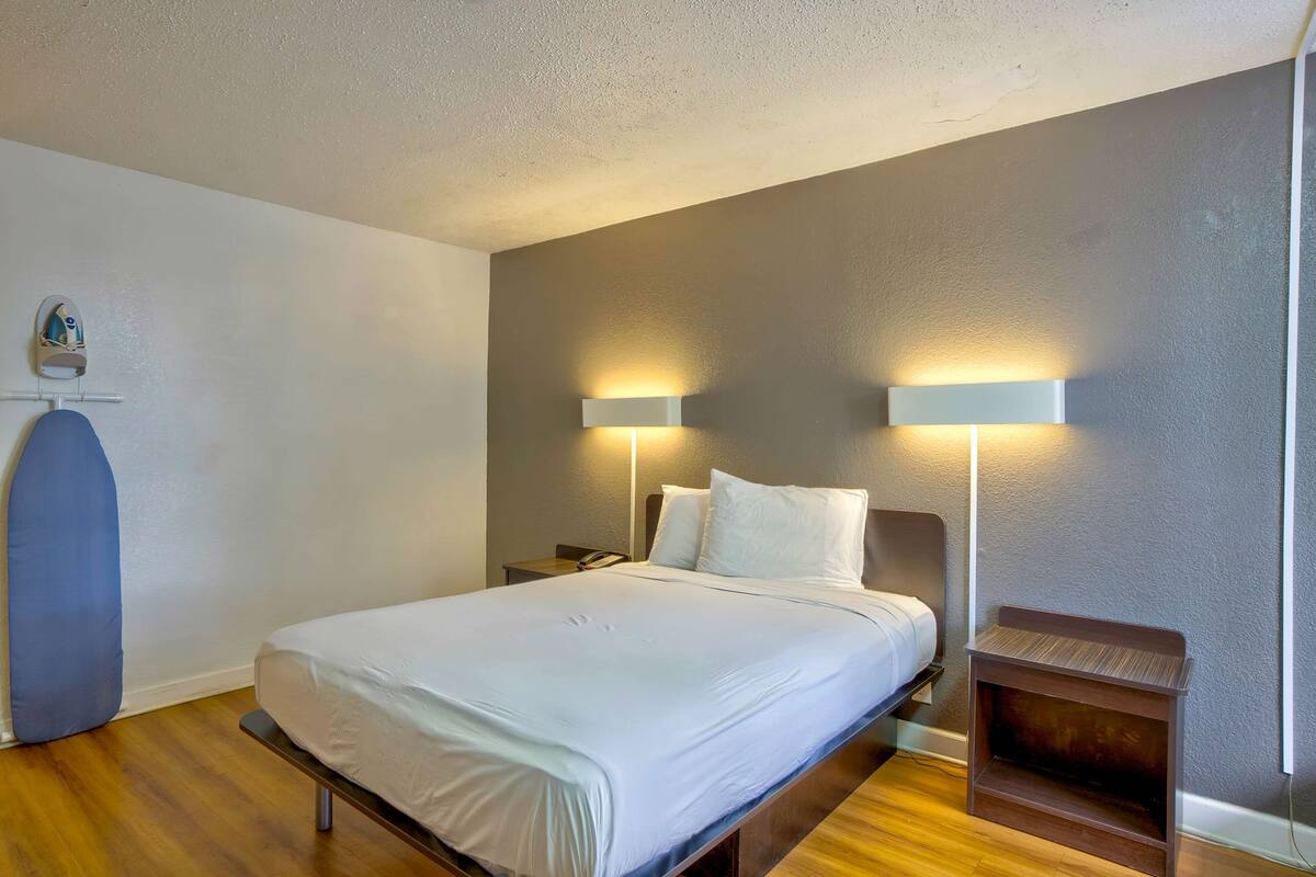 A neatly made bed in a hotel room with two bedside lamps, a small wooden nightstand, and a wall-mounted ironing board.