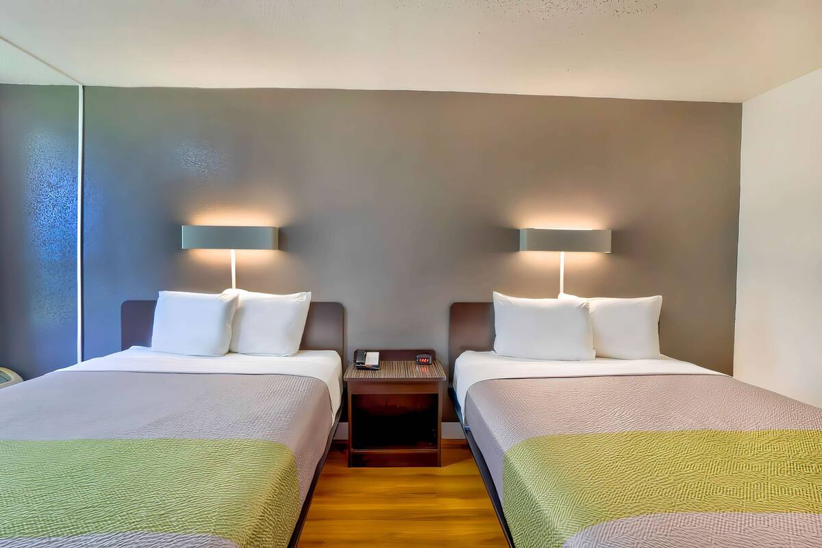 A hotel room with two beds, a nightstand with a phone and clock, and wall-mounted lamps.
