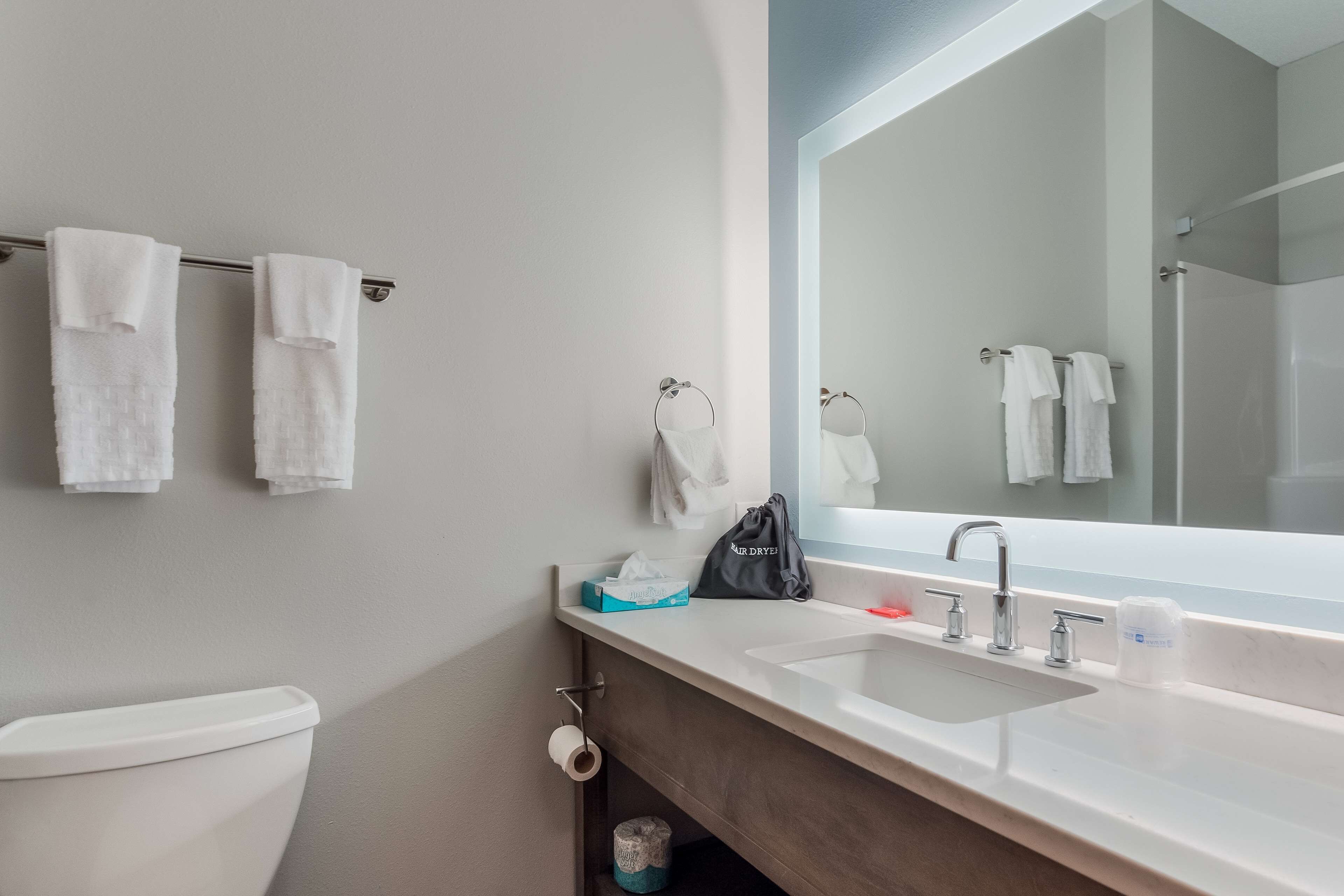 Un baño moderno con inodoro, lavamanos y un espejo grande. Hay toallas colgadas en un perchero y un bolso sobre la encimera.