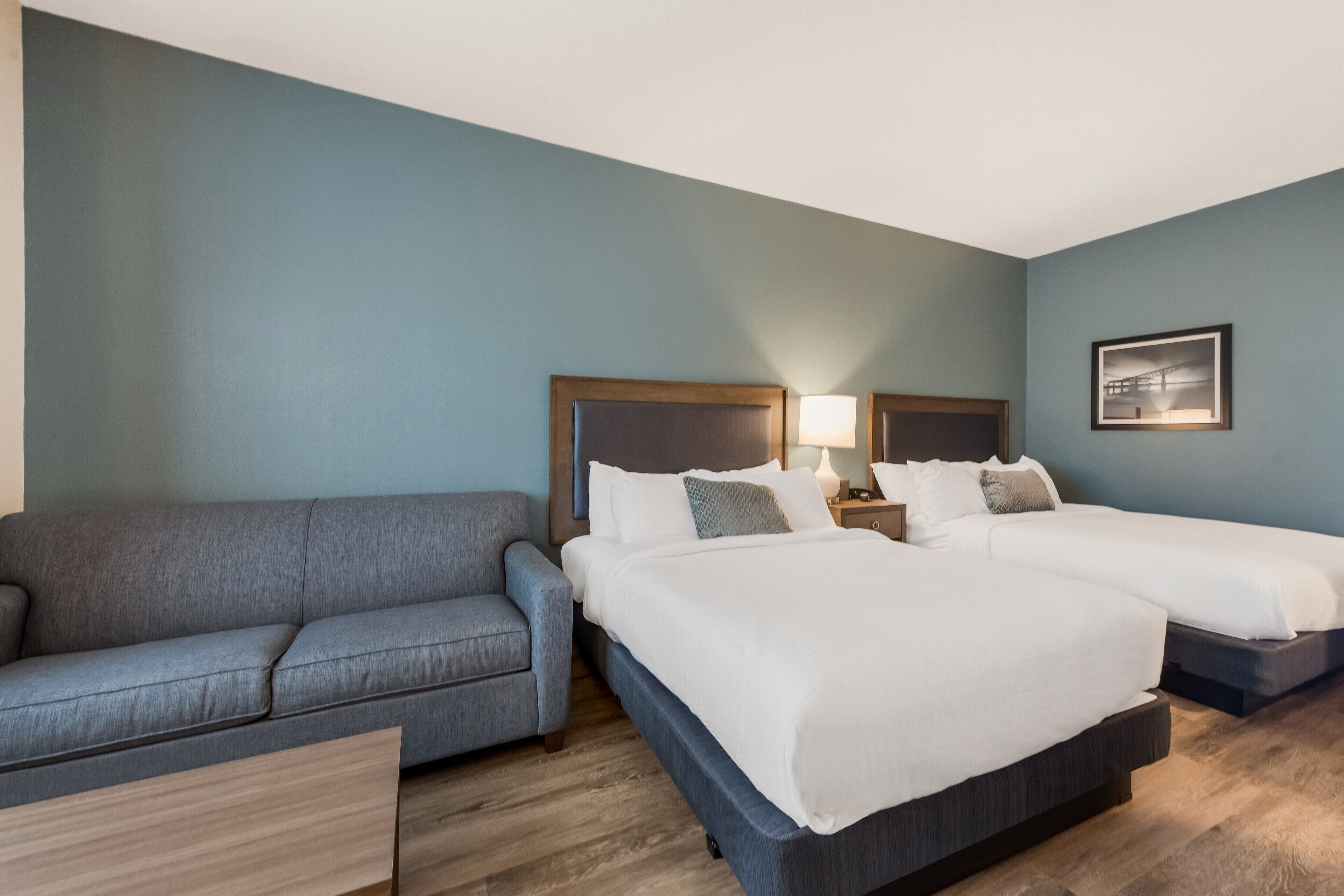 Une chambre d’hôtel avec un canapé gris, deux lits simples, une table de chevet en bois avec une lampe et un tableau encadré au mur.