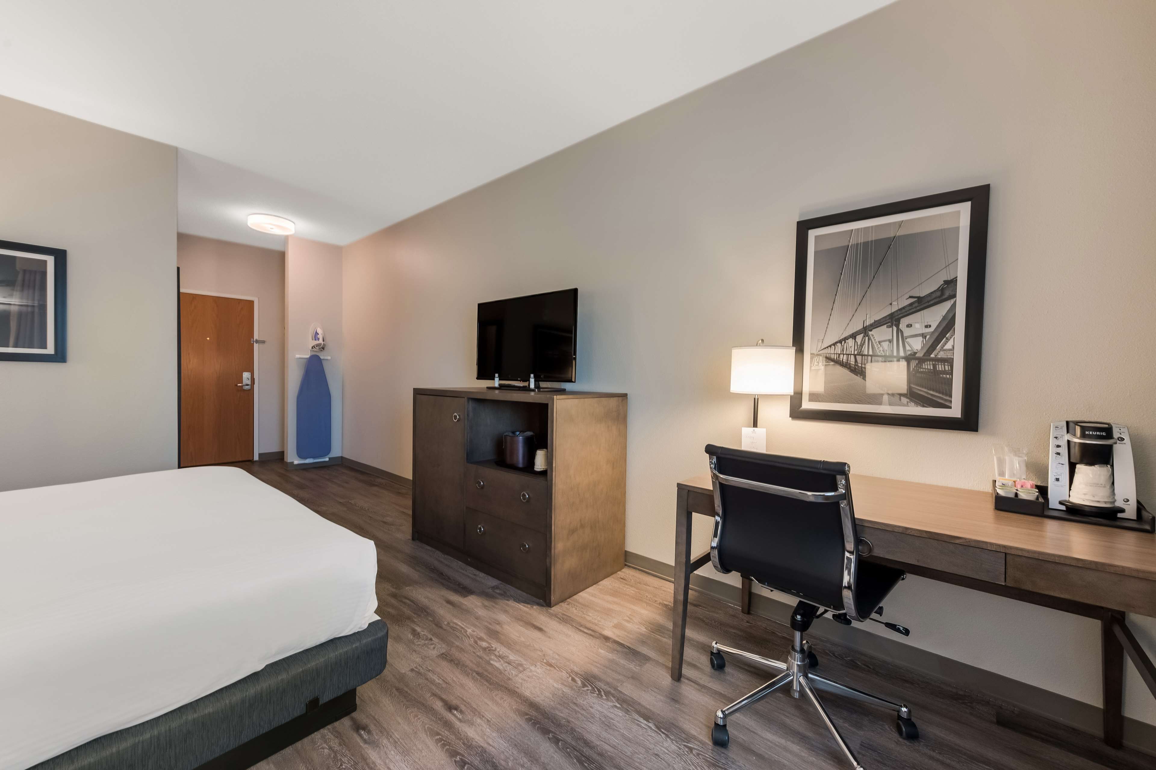 Habitación de hotel con cama, escritorio, silla, cafetera, televisión y un cuadro en la pared.