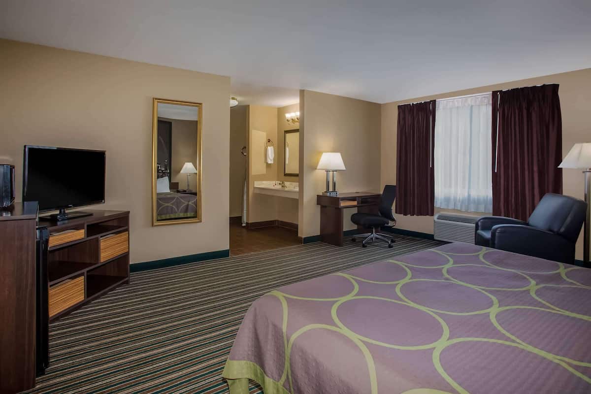 A hotel room with a large bed, a desk with a chair, a television, a bathroom with a sink and mirror, and a window with curtains.