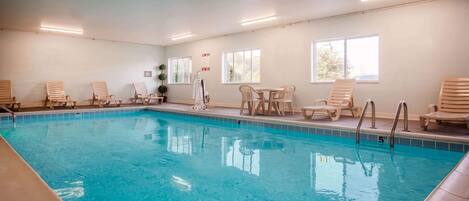 An indoor swimming pool with clear water, surrounded by wooden lounge chairs and a table.