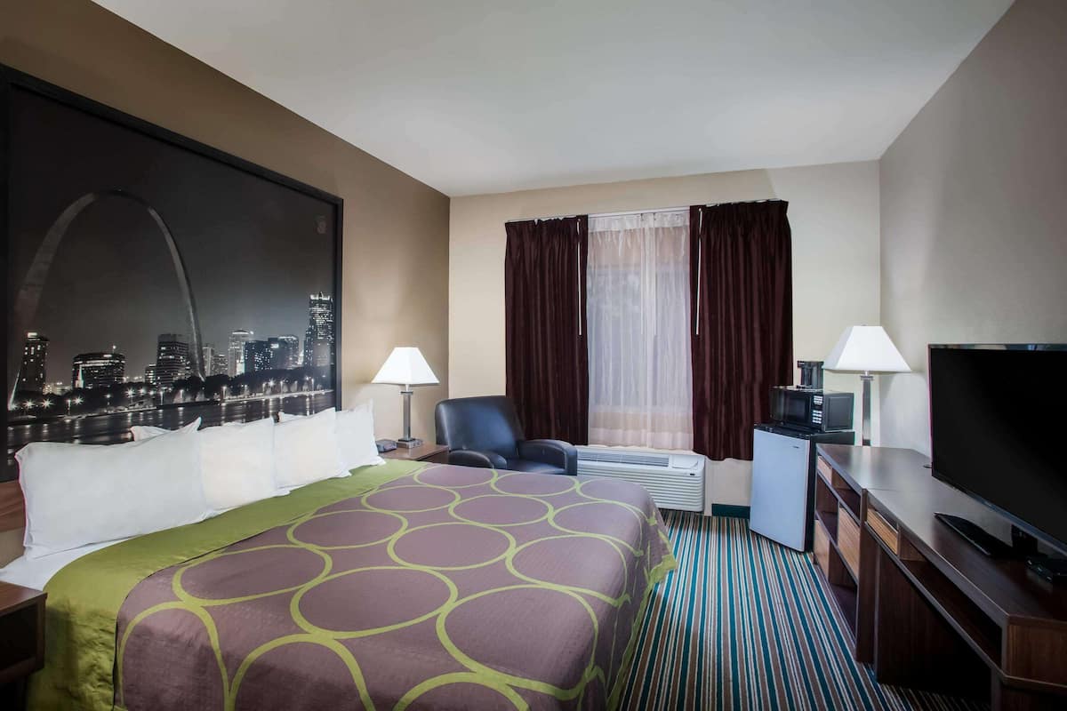 A hotel room with a large bed, two chairs, a desk with a TV, a mini-fridge, and a cityscape view.