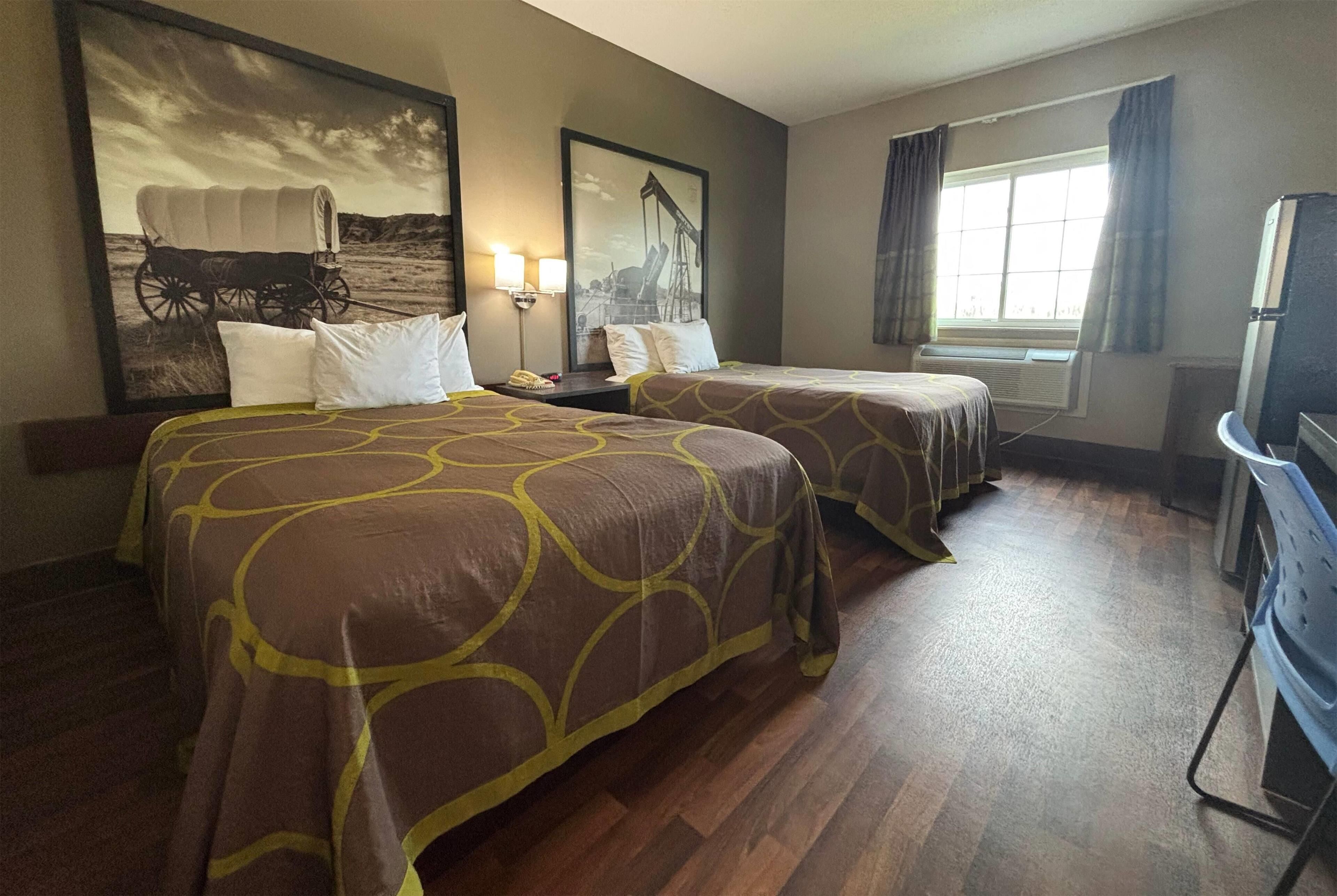 A hotel room with two beds, a large painting of a covered wagon, a desk with a lamp, and a window with curtains.