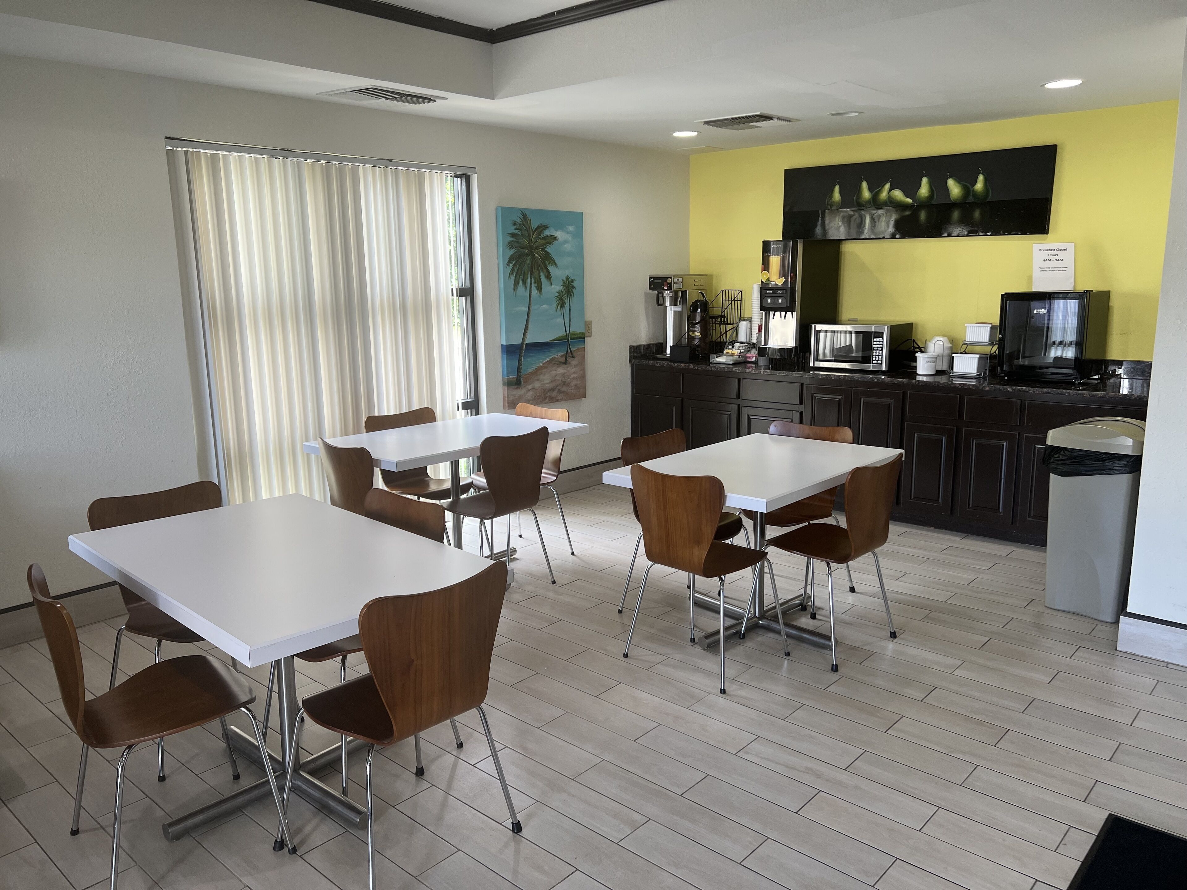Uma sala de jantar moderna com mesas brancas e cadeiras castanhas, uma cozinha com armários e eletrodomésticos escuros, e uma parede amarela que destaca o ambiente.