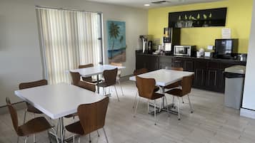 Uma sala de jantar moderna com mesas brancas e cadeiras castanhas, uma cozinha com armários e eletrodomésticos escuros, e uma parede amarela que destaca o ambiente.