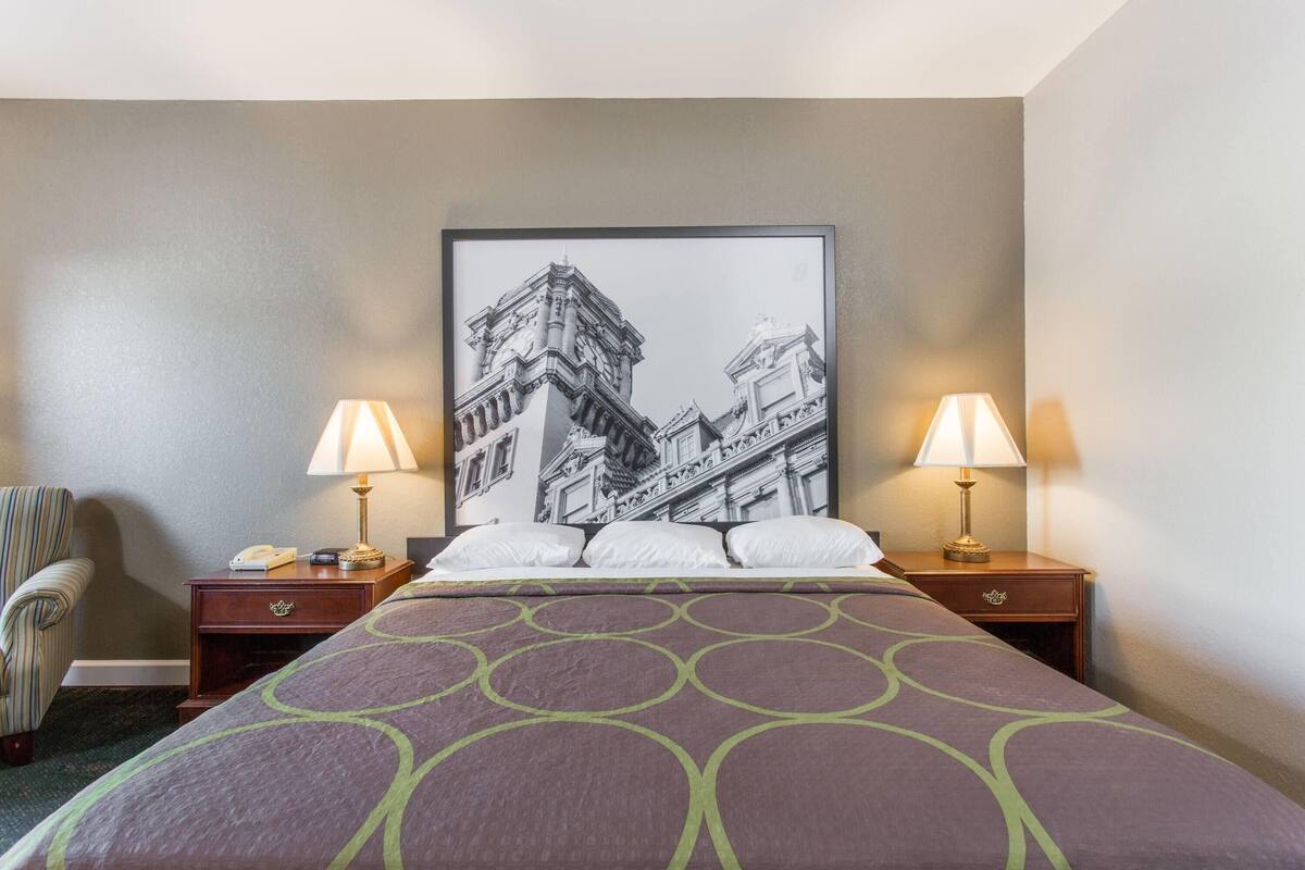 A hotel room with a bed, two bedside tables with lamps, a chair, and a large framed picture on the wall.