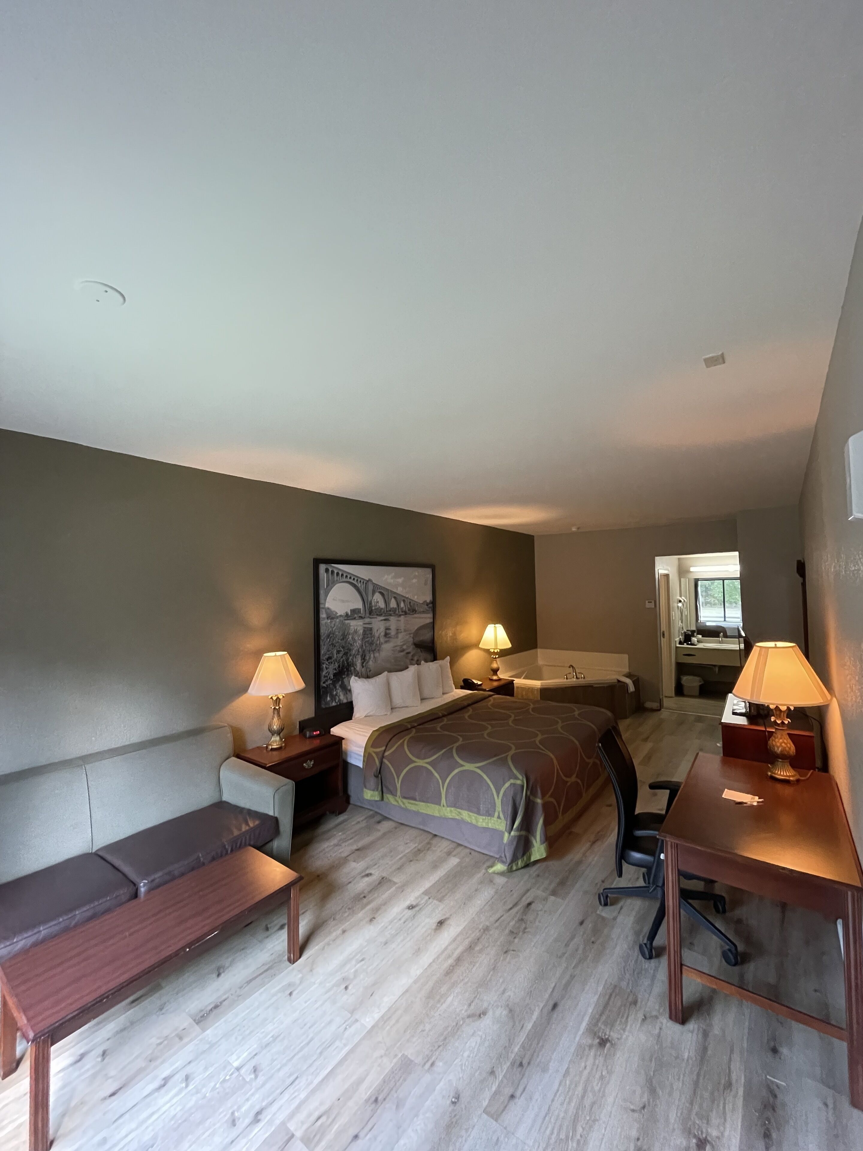Une chambre d’hôtel comprenant un lit, un bureau, une chaise, un banc, une table de chevet, une lampe et une salle de bain visible à travers une porte ouverte.