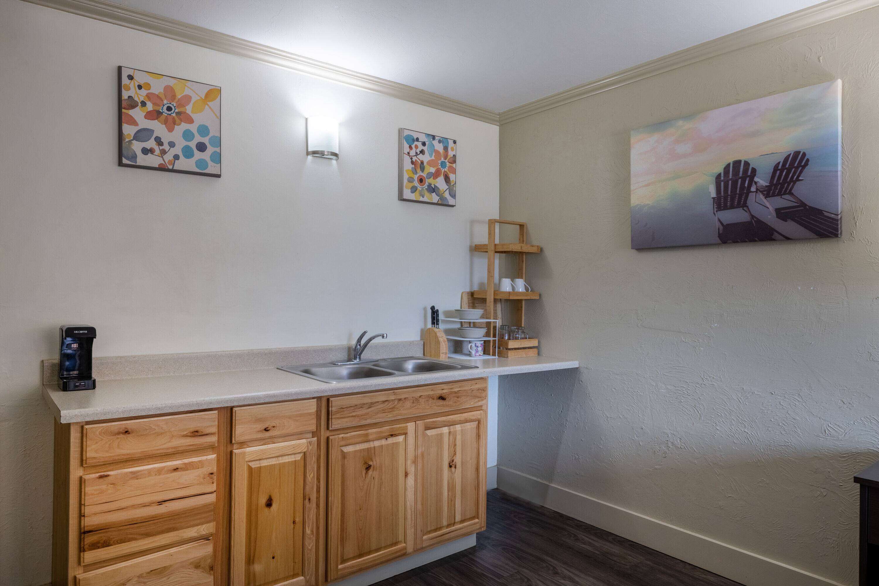 A kitchen with wooden cabinets, a countertop, a sink, and a coffee maker. There are three wall-mounted artworks and a shelf with cups and bowls.