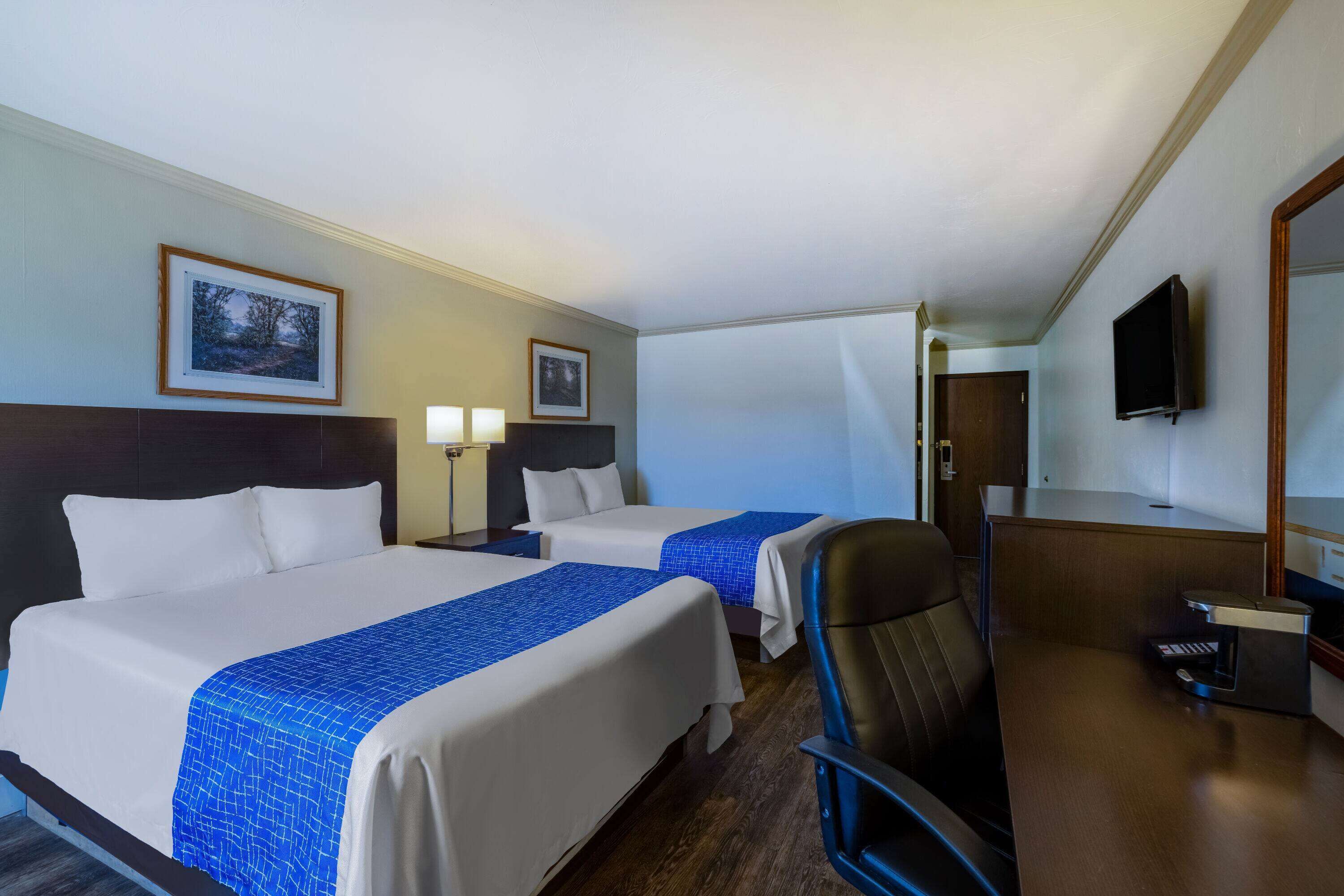 Une chambre d’hôtel avec deux lits, un bureau, une chaise, une télévision et des tableaux encadrés au mur.