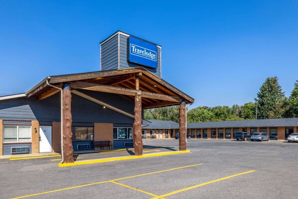 A Travelodge with a covered parking area, a brick facade, and a row of cars parked in the lot. - Travelodge by Wyndham Livingston Yellowstone (Livingston)