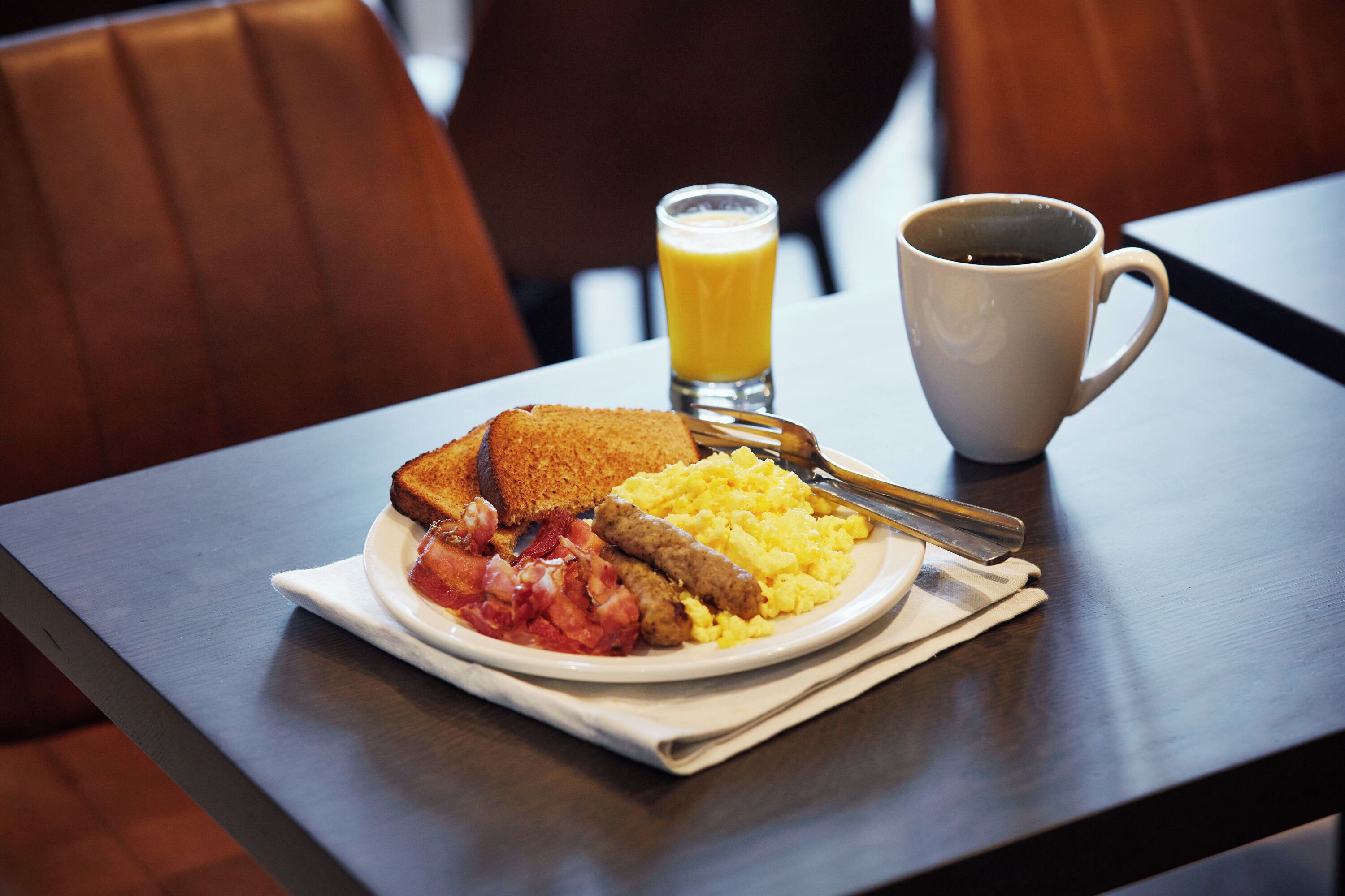 A breakfast plate with toast, bacon, scrambled eggs, and a glass of orange juice on a table.