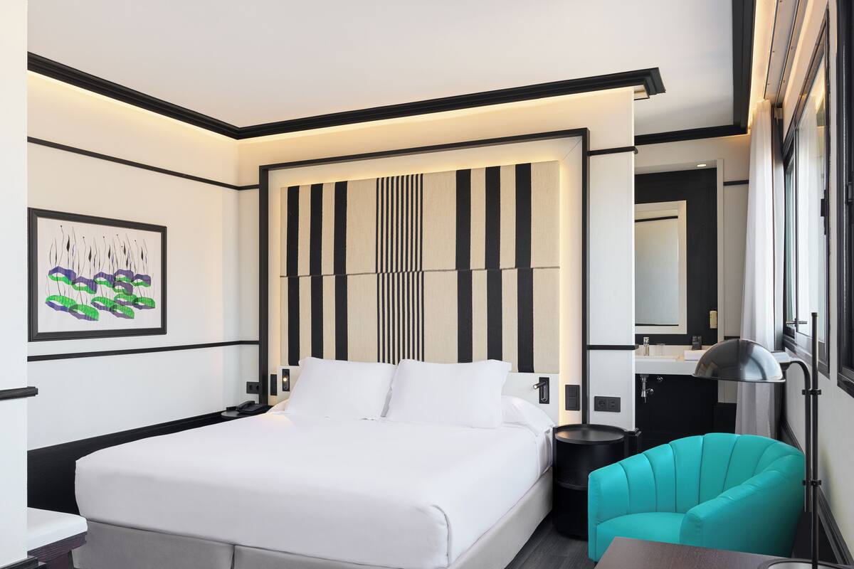 Ein modernes Hotelzimmer mit einem großen Bett, einem Schreibtisch, einem Stuhl, einem Badezimmer mit Spiegel und einem an der Wand hängenden Kunstwerk.